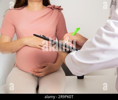 Una ragazza incinta viene esaminata da un medico generico che scrive una lista di prescrizioni per la nutrizione di una donna incinta. Foto Stock