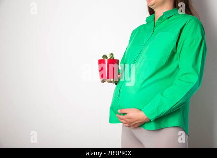 Una ragazza incinta in una camicetta verde tiene un piatto di cactus rosso. Coznept di dolori da taglio e coliche nell'addome nelle donne incinte. Cistite e patologia Foto Stock