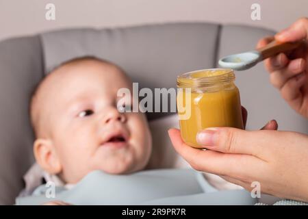 La madre tiene un barattolo di purea vegetale sullo sfondo di un bambino di 6 mesi. Il primo cibo per bambini. Foto Stock