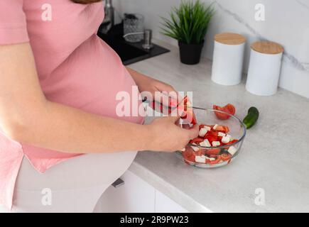 Una ragazza incinta prepara un'insalata di verdure di cetrioli e pomodori. Il concetto di nutrizione delle donne incinte con vitamine e macronutrienti in v Foto Stock