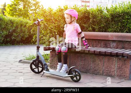 Una bella bambina di sette anni siede su una panchina con un equipaggiamento di protezione rosa accanto a uno scooter. Il concetto di ciclismo e scootering senza infortuni. Foto Stock