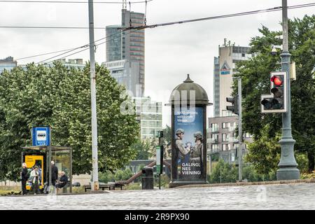 Banner pubblicitario che informa sul vertice NATO 2023 nel centro di Vilnius, capitale della Lituania, con persone in attesa alla fermata dell'autobus e traffico rosso Foto Stock