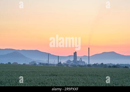 Panorama industriale con camini e torri di fabbrica di calcestruzzo in campi di grano verde e montagne all'alba. Emissioni di gas da fumo. Foto Stock