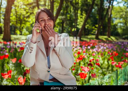 Giovane donna d'affari caucasica che parla al telefono tenendo i notebook nel parco primaverile. Imprenditore che fa una pausa all'aperto e si rilassa nel giardino pubblico Foto Stock