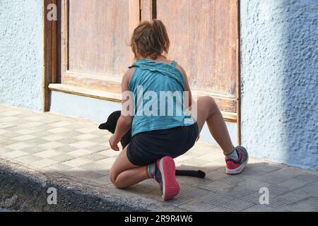 Una ragazza inginocchiata irriconoscibile con un gatto nero davanti a lei in una strada soleggiata. Foto Stock