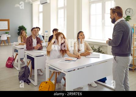 Gli studenti seduti alle scrivanie alzano le mani per rispondere alla domanda del loro insegnante Foto Stock