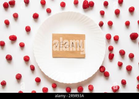 La parola vegan è scritta su carta e giace su un piatto su uno sfondo di lamponi sparsi su uno sfondo bianco, senza carne, frutta Foto Stock