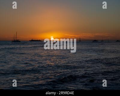 Il tramonto sull'Oceano Pacifico visto da una spiaggia di Maui brilla di arancione e mette in evidenza le onde e le navi di passaggio Foto Stock