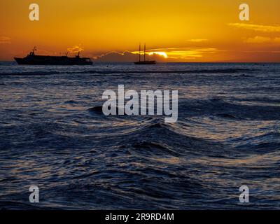 Il tramonto sull'Oceano Pacifico visto da una spiaggia di Maui brilla di arancione e mette in evidenza le onde e le navi di passaggio Foto Stock