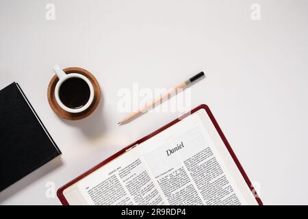 Lastra piatta della Bibbia cristiana aperta al Libro di Daniele con caffè espresso, taccuino e matita su sfondo bianco foto di scorta Foto Stock