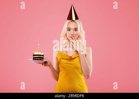 Felice ragazza con un cappello da festa che tiene un pezzo di torta e chiude la bocca con la mano, festeggia il compleanno, sfondo rosa Foto Stock