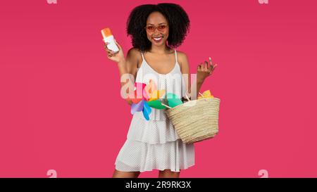 Felice giovane donna riccia nera con occhiali da sole con cestino mostra barattolo crema solare Foto Stock