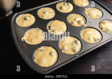 Muffin in stagno foderato con involtini di carta e ripieno con impasto di muffin ai mirtilli: Impasto di muffin ai mirtilli non cotto in una padella con attrezzi da cucina Foto Stock
