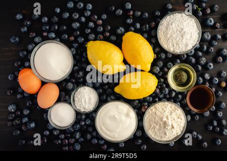 Muffin ai mirtilli con glassa al limone su sfondo di legno scuro: Mirtilli freschi, limoni e altri ingredienti crudi su uno sfondo rustico di legno Foto Stock