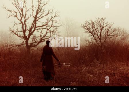 Donna vittoriana che cammina con la lanterna attraverso paludi boscose invernali. La nebbia nebulizzata evoca un altro mondo e un'altra volta. Foto Stock