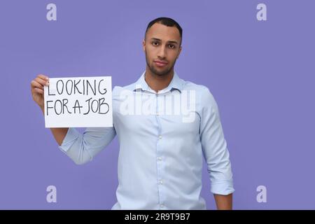 Giovane disoccupato con un cartello con una frase che cerca Un lavoro su sfondo violetto Foto Stock