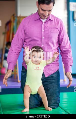 Bella bambina di dieci mesi che impara a camminare aiutata da suo padre. Concetto di stimolazione precoce per i bambini piccoli. Foto Stock