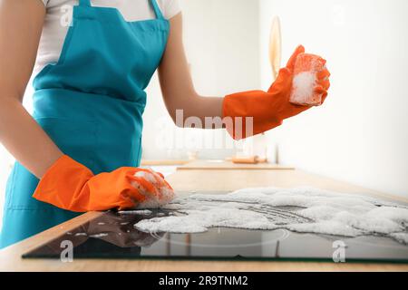 Donna con guanti di gomma arancione che pulisce la stufa elettrica con spugna Foto Stock