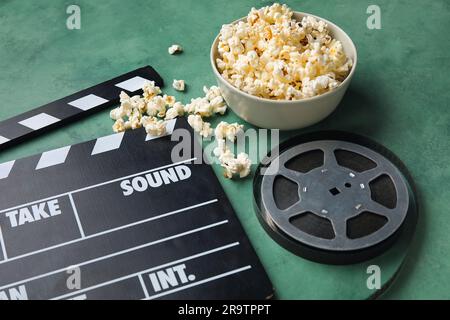 Ciotola con gustosi popcorn, clapperboard e bobina di pellicola su sfondo verde Foto Stock