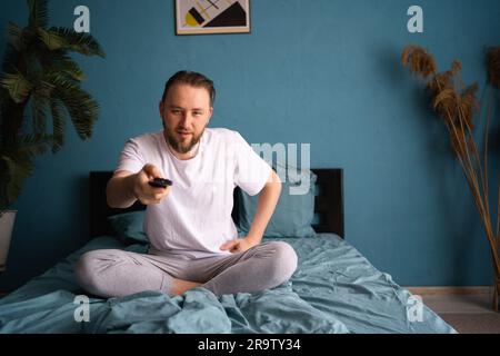 Un uomo insonne in pigiama che guarda la tv mentre è seduto sul letto con telecomando Foto Stock