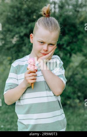 Un bambino triste stringeva la guancia con la mano e teneva un cono gelato nell'altra mano. Problemi di sensibilità dei denti in un bambino o di decadimento dei denti. Foto Stock