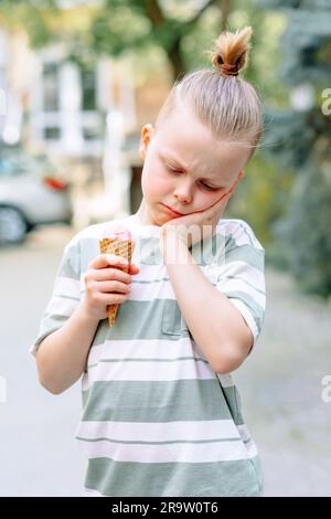 Un bambino triste stringeva la guancia con la mano e teneva un cono gelato nell'altra mano. Problemi di sensibilità dei denti in un bambino o di decadimento dei denti. Foto Stock