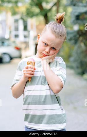 Un bambino triste stringeva la guancia con la mano e teneva un cono gelato nell'altra mano. Problemi di sensibilità dei denti in un bambino o di decadimento dei denti. Foto Stock