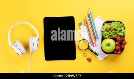 Tablet con schermo vuoto, forniture scolastiche, cancelleria e cestino per il pranzo su sfondo giallo. Educazione e nutrizione. Posa piatta con spazio vuoto per il Foto Stock