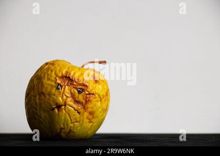 Una mela marcio e lenta con un triste sorriso dipinto su uno sfondo bianco su un tavolo nero, di cattivo umore Foto Stock