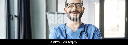 ritratto di giovane e gioioso operatore sanitario con barba, occhiali e stetoscopio guardando la macchina fotografica in clinica moderna, seminario di formazione di pronto soccorso a Foto Stock