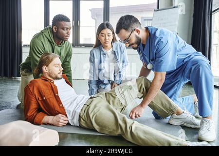 seminario di pronto soccorso, istruttore medico in occhiali da vista e uniforme applicazione di benda compressione sulla gamba dell'uomo vicino donna asiatica e afro americano, impo Foto Stock