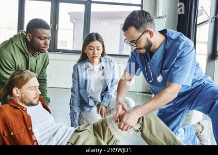 operatore sanitario professionale che applica la benda di compressione sulla gamba dell'uomo vicino a studenti multietnici durante il seminario di primo soccorso in sala di formazione, importa Foto Stock