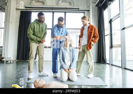 Uomo multietnico che guarda la testa di fasciatura paramedica professionale della donna asiatica vicino al manichino RCP durante il seminario di formazione di primo soccorso, importanza di emer Foto Stock