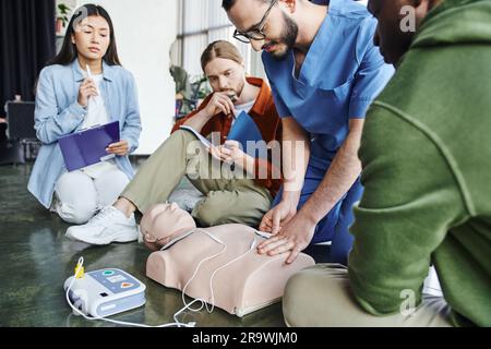 Seminario di primo soccorso, istruttore medico che applica gli elettrodi per defibrillazione sul manichino RCP vicino a team multietnico con clipboard e notebook nella sala di addestramento, h Foto Stock