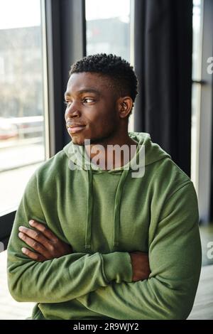 uomo d'affari afro-americano con felpa verde con cappuccio, con braccia piegate e guardando lontano vicino alla finestra in un ufficio moderno, giovane imprenditore Foto Stock
