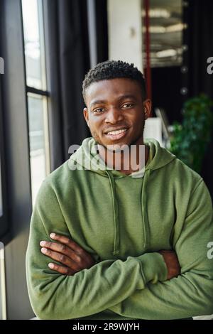 giovane e carismatico imprenditore afro-americano con felpa verde e alla moda in piedi con le braccia piegate e sorridendo alla macchina fotografica, emozione felice, cont Foto Stock