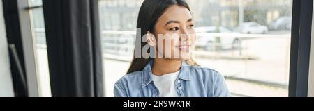 attraente e gioiosa donna asiatica con camicia in denim blu, con capelli brunette in piedi vicino alla finestra in un ufficio moderno e guardando lontano, giovane e di successo Foto Stock