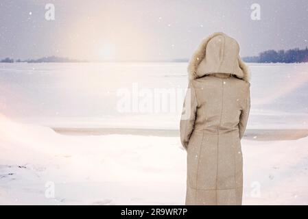 Una donna con un cappotto di pelle con cappuccio di pelliccia sta guardando il fiume Dnieper ghiacciato durante una fredda e triste mattinata invernale sotto la neve Foto Stock