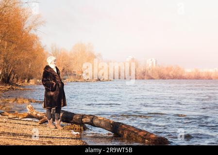 Una donna matura che indossa una calda pelliccia e un cappello di lana resta vicino al fiume Dnieper a Kiev, Ucraina, durante la stagione invernale Foto Stock