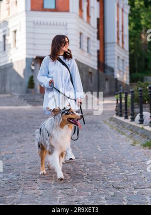 Giovane donna e il suo cane pastore asutraliano al guinzaglio, attenzione selettiva sul cane. Camminare con gli animali domestici in centro città, immagine di stile di vita Foto Stock