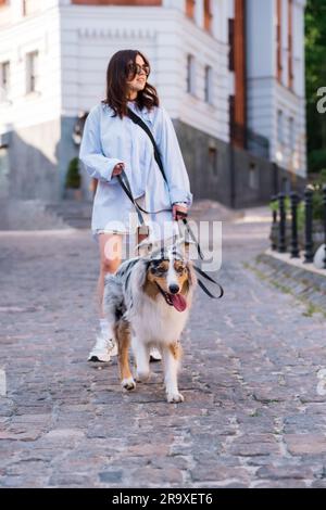 Giovane donna e il suo cane pastore asutraliano al guinzaglio, attenzione selettiva sul cane. Camminare con gli animali domestici in centro città, immagine di stile di vita Foto Stock