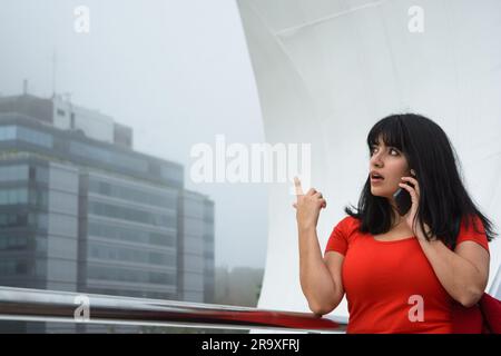 La giovane donna latina di etnia venezuelana, vestita di rosso, è sorpresa all'aperto a parlare al telefono e a puntare il dito verso l'alto, in piedi sulla PUE Foto Stock
