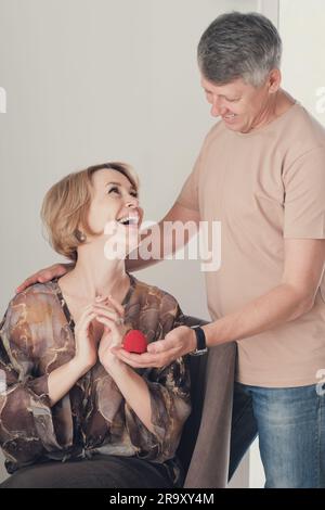 un uomo sorridente di mezza età regala un regalo a una donna eccitata. Il marito fa un regalo per sua moglie, un gioiello in una confezione regalo di velluto rosso. Shot verticale. Foto Stock