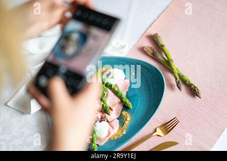 Una donna che scatta una foto del cibo con uno smartphone. Antipasto con prosciutto al rosmarino, asparagi, arachidi e taleggio sul piatto blu. Foto Stock