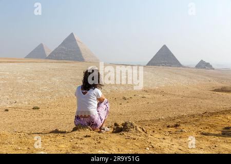 Una vista posteriore di una giovane donna seduta sulla sabbia guardando le famose piramidi di Giza in Egitto Foto Stock