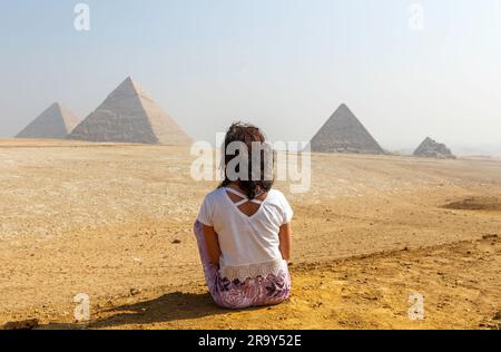 Una vista posteriore di una giovane donna seduta sulla sabbia guardando le famose piramidi di Giza in Egitto Foto Stock