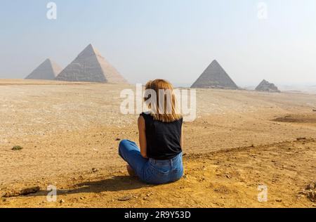 Una vista posteriore di una giovane donna seduta sulla sabbia guardando le famose piramidi di Giza in Egitto Foto Stock