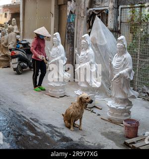 Saigon, Vietnam, 23 marzo 2023. Un cane attende pazientemente l'attenzione mentre una donna vietnamita dipinge statue di Buddha su una strada laterale a Saigon. Foto Stock