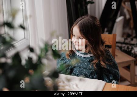 Adorabile bambina con i capelli ricci indossa un abito in stile rurale festoso sorride, seduta accanto al tavolo e sostiene il mento con la mano a casa. Felice c Foto Stock