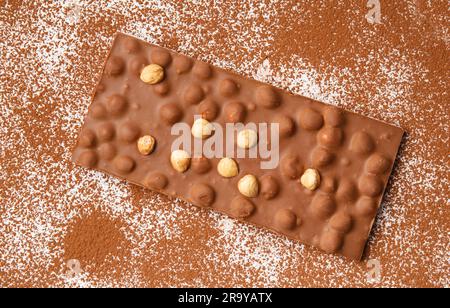 Il cioccolato al latte con nocciole si trova su uno sfondo bianco cosparso di cacao in polvere dall'alto. Fondo di cacao in polvere e cioccolato. Foto Stock